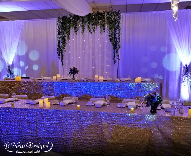 A gorgeous white flowy backdrop lit up with blueish uplighting, the center panel lined with full draping greenery, twinkle lights peeking through the draping chiffon. The end panels cinched in the middle with swags of hanging eucalyptus.